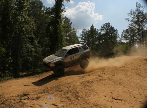 1999 Isuzu Vehicross - An SUV navigates a dirt track, its tires kicking up a cloud of dust under a partly cloudy sky.