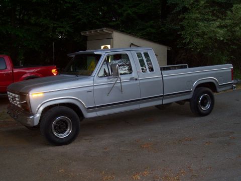 1978 Ford F-150 - A silver pickup truck with a crew cab and long bed is parked outside near some trees and a small building.