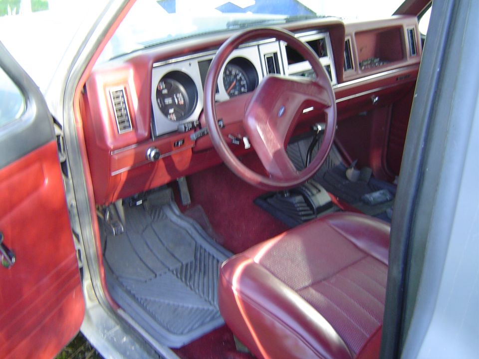 Red Ford interior through open driver's door showing dash gauges, red steering wheel, and floor-console shifter
