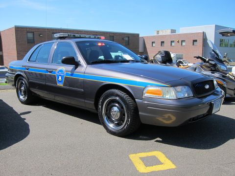 1991 Ford LTD Crown Victoria - Gray Ford Crown Victoria police car with a blue and yellow body stripe and a roof light bar, parked beside a motorcycle.