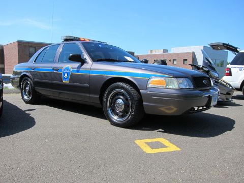 1991 Ford LTD Crown Victoria - Gray Ford Crown Victoria police sedan with a blue and yellow stripe and roof light bar parked over a yellow lot marking.