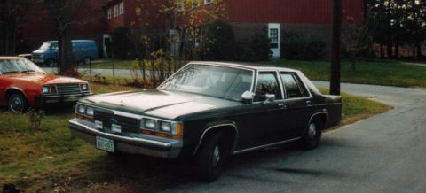1991 Ford LTD Crown Victoria - Front three-quarter driver-side view of a dark green Ford LTD Crown Victoria sedan parked on a driveway beside a red barn-style building, w…