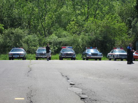 1991 Ford LTD Crown Victoria - Five older full-size police sedans parked side by side along a wooded lot edge as several people stand nearby.