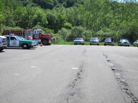 1991 Ford LTD Crown Victoria - broad cracked parking lot holds a green-and-white police car, a red fire engine, and a distant row of vintage sedans.