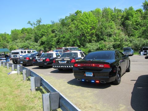 1991 Ford LTD Crown Victoria - black Dodge Charger police sedan parked at the end of a row of marked cruisers and an ambulance beside a guardrail.