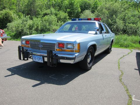 1991 Ford LTD Crown Victoria - light-blue 1980s Ford LTD Crown Victoria police sedan with front push bar and roof light bar parked on pavement.