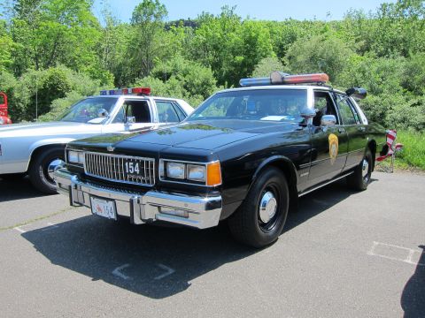 1991 Ford LTD Crown Victoria - black 1980s Chevrolet Caprice police sedan marked 154 with roof light bar and chrome bumper parked in a lot.