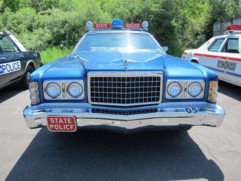 1991 Ford LTD Crown Victoria - Head-on view of a blue 1970s Ford LTD marked STATE POLICE with chrome grille, dual round headlights and roof beacons.