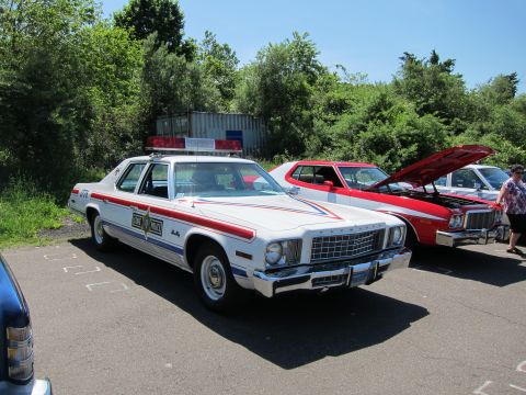 1991 Ford LTD Crown Victoria - white 1970s Plymouth Fury state-police sedan with red-and-blue striping parked beside a red car at a show.
