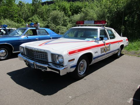 1991 Ford LTD Crown Victoria - white 1970s Plymouth Fury marked STATE POLICE with red striping and roof light bar parked beside a blue cruiser.