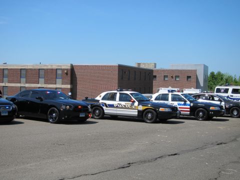 1991 Ford LTD Crown Victoria - lineup of police vehicles including a black Dodge Charger and white sedans parked in front of a brick building.