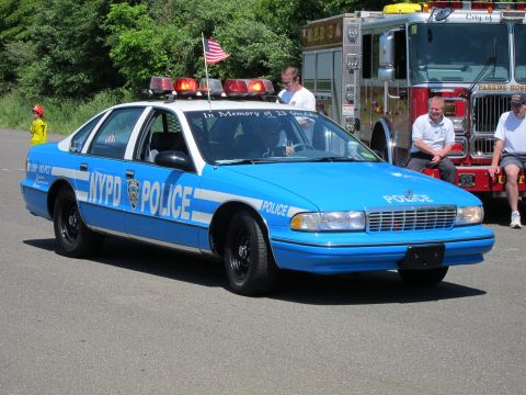 1991 Ford LTD Crown Victoria - blue NYPD Caprice police car with roof lightbar and American flag parked beside a red fire engine at a show.
