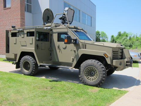 1991 Ford LTD Crown Victoria - military-style olive-green armored vehicle with a roof antenna dish and turret, parked on pavement by a building.