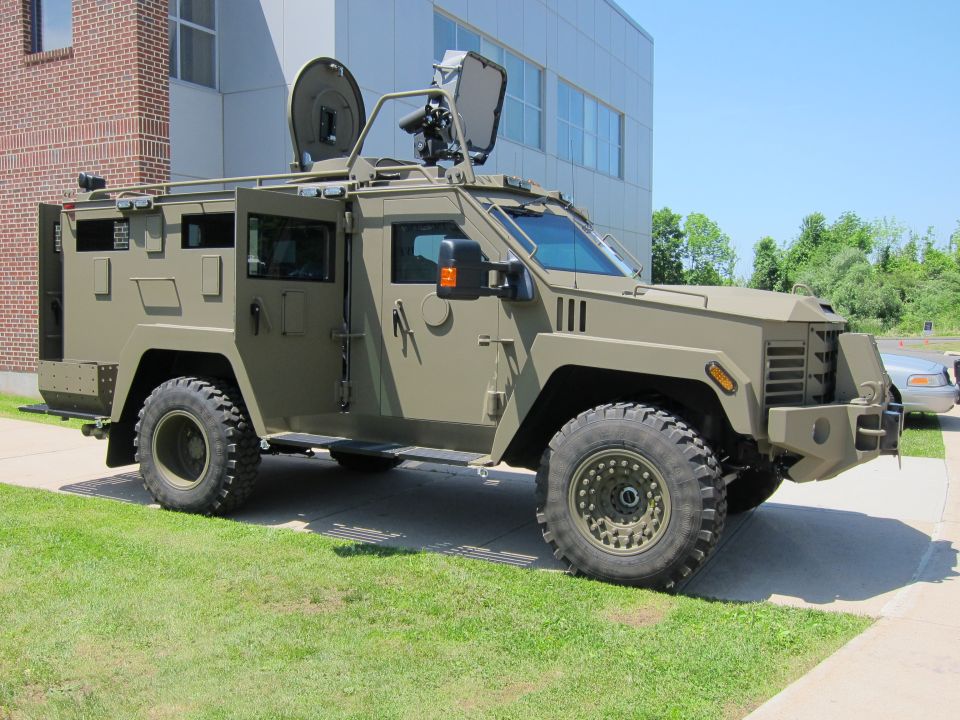 Olive-green armored truck with antenna mast and gun turret parked beside a building
