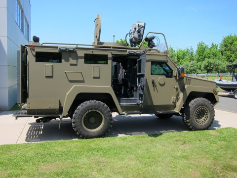 1991 Ford LTD Crown Victoria - Profile of a tan-green armored tactical vehicle with an open crew door and roof-mounted camera mast, on a paved walkway.