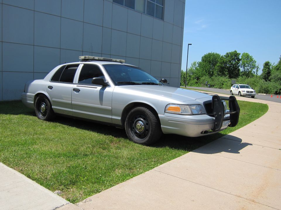 Silver Ford Crown Victoria police sedan with push bar parked on grass by a building