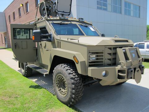 1991 Ford LTD Crown Victoria - Three-quarter front view of a tan Lenco armored tactical vehicle parked on grass beside a brick and metal building.
