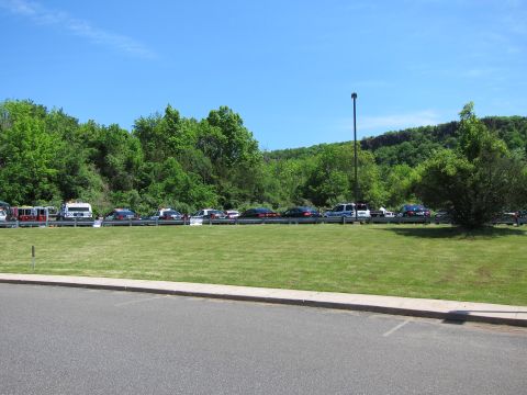 1991 Ford LTD Crown Victoria - wide view of a grassy field with a distant row of parked cars and trucks along a guardrail, backed by a wooded hill.