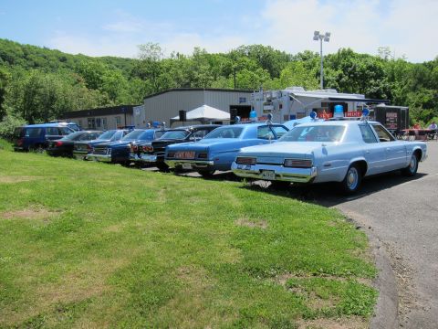 1991 Ford LTD Crown Victoria - lineup of vintage 1970s blue State Police sedans parked diagonally on grass, with tents and a building in the background.