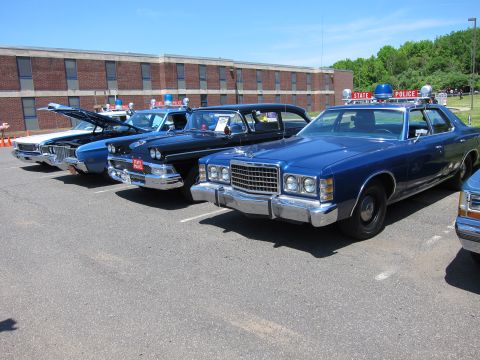 1991 Ford LTD Crown Victoria - lineup of restored 1970s blue Ford State Police sedans parked diagonally in front of a long brick building.