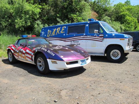 1991 Ford LTD Crown Victoria - flag-themed Connecticut State Police Corvette sports car beside a stars-and-stripes D.A.R.E. Ford van on a gravel lot.