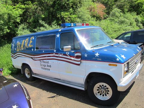 1991 Ford LTD Crown Victoria - Side view of a blue and white Ford van decorated with stars, a flag stripe and gold D.A.R.E. lettering parked on gravel.