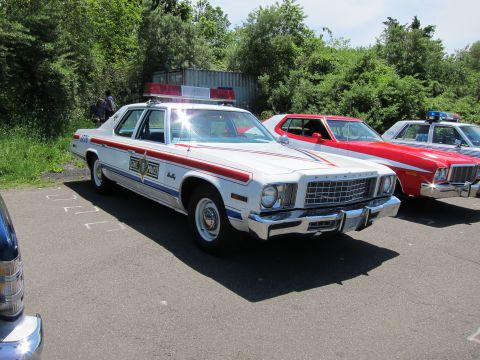 1991 Ford LTD Crown Victoria - white 1970s Plymouth full-size police sedan with a red side stripe and roof light bar parked beside other show cars.