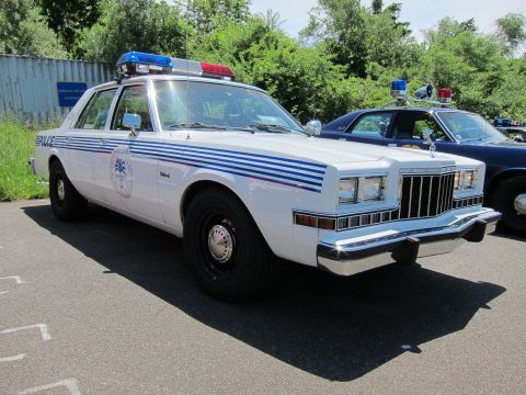 1991 Ford LTD Crown Victoria - white 1980s Dodge police sedan with blue striping, a door seal emblem and a red and blue roof light bar on asphalt.