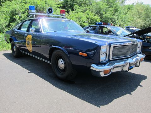 1991 Ford LTD Crown Victoria - dark blue 1970s Plymouth police sedan with a gold door seal, spotlight and roof light bar parked at a car show.