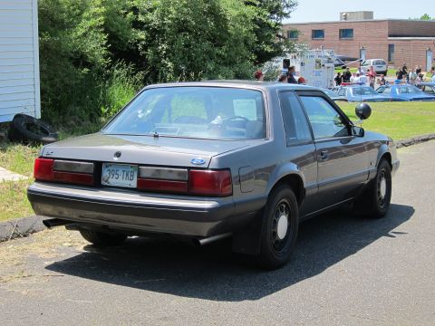 1991 Ford LTD Crown Victoria - Rear view of a gray Fox-body Ford Mustang notchback coupe with plate 395-TKB parked near a grassy lot and brick building.