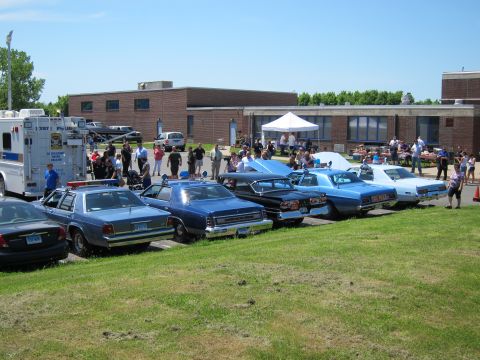 1991 Ford LTD Crown Victoria - lineup of classic police sedans and a box truck parked on grass with a crowd and brick building behind at a car show.