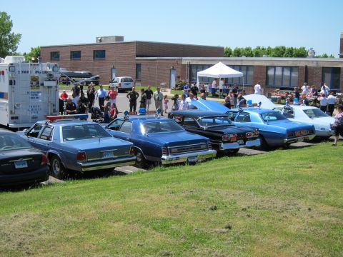 1991 Ford LTD Crown Victoria - row of vintage police sedans parked on grass beside a command box truck with spectators near a brick building.