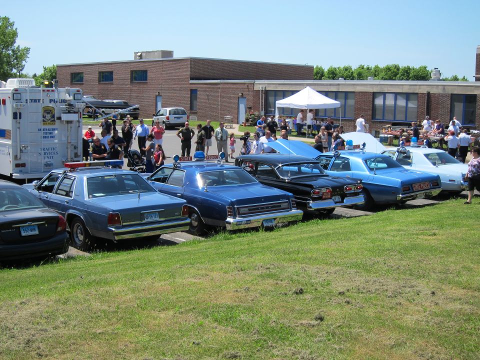 Lineup of vintage police sedans on grass with crowd and box truck behind