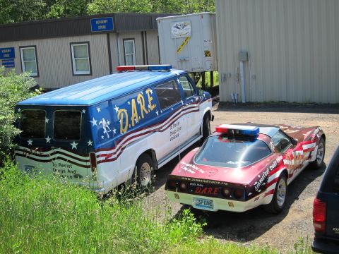 1991 Ford LTD Crown Victoria - stars-and-stripes D.A.R.E. Ford van and a flag-painted Corvette parked on gravel beside an Academy Store building.