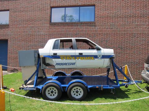 1991 Ford LTD Crown Victoria - white Connecticut State Police rollover-simulator car body sits on a blue tandem-axle trailer in front of a brick building.