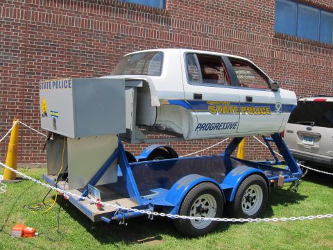 1991 Ford LTD Crown Victoria - Three-quarter rear view of a white Connecticut State Police rollover-demonstration car body on a blue trailer beside a brick wall.