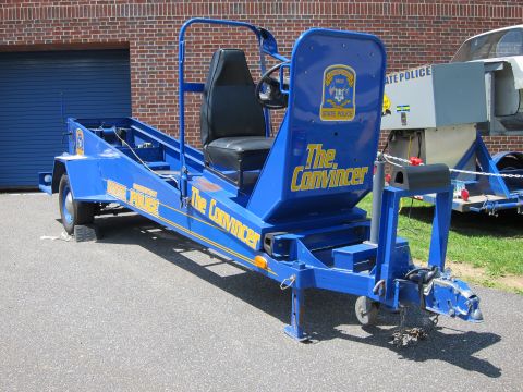 1991 Ford LTD Crown Victoria - blue State Police seatbelt-demonstration trailer labeled The Convincer with a single seat and steering wheel sits on pavement.