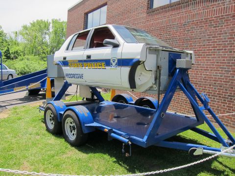 1991 Ford LTD Crown Victoria - white Connecticut State Police rollover-simulator car body tilted on a blue trailer on grass beside a brick building.