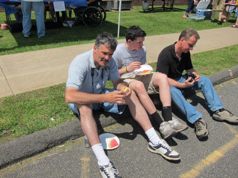 1991 Ford LTD Crown Victoria - Three men sit on a curb eating slices of watermelon from paper plates at a sunny outdoor gathering.