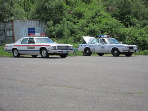 1991 Ford LTD Crown Victoria - Two vintage police cars, a white sedan with red stripes and number 1776 and a gray cruiser, parked on a gravel lot near trees.
