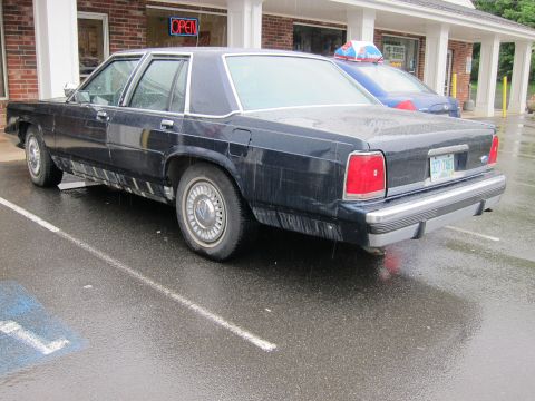 1991 Ford Crown Victoria LTD - A dark sedan is parked in a handicapped spot on a rainy day, with another vehicle visible behind it.