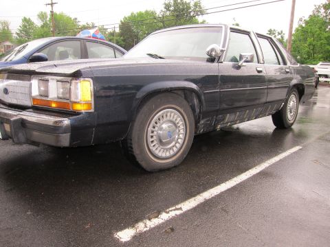 1991 Ford Crown Victoria LTD - A dark blue sedan is parked on a wet asphalt surface, with another vehicle visible behind it.