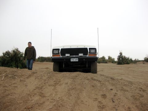1979 Ford Bronco - A man walks past a white Ford truck parked on a dirt mound under a hazy sky.
