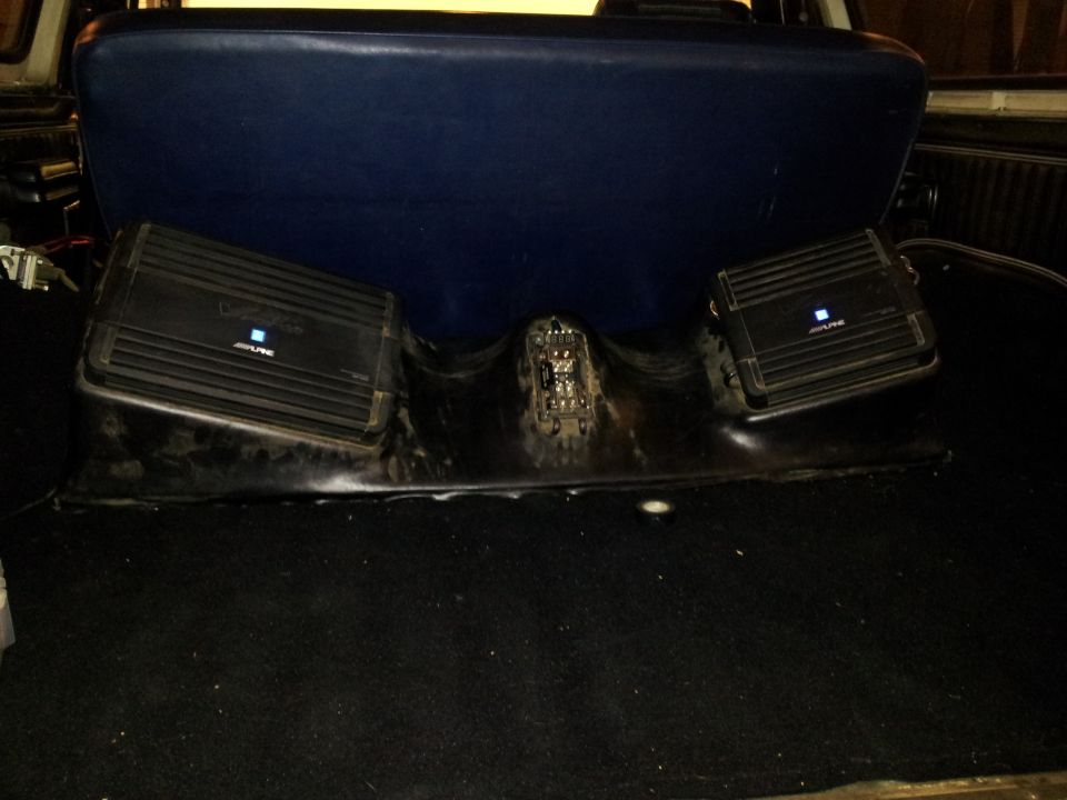 Vehicle interior with two black Alpine car amplifiers mounted on a black plastic housing behind a blue seat