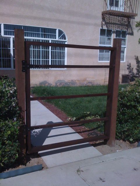 1968 Ford Mustang - A rusty metal gate stands open on a sidewalk, with a grassy area and building behind it.