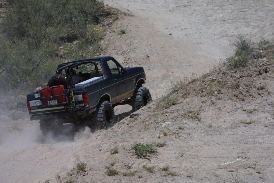 Dark blue topless lifted Ford Bronco climbing dusty desert hill with fuel cans on rear rack