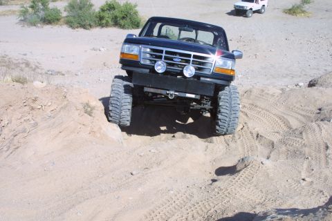 1994 Ford Bronco - A lifted black Ford F-Series truck navigates a sandy, off-road terrain with a white SUV visible in the distance.