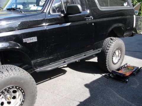 1993 Ford Bronco - Side profile of a black Ford Bronco full-size SUV, lifted with large off-road tires on chrome beadlock-style wheels and a black tubular sid…