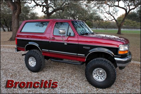 1993 Ford Bronco - Two-tone red and black lifted Ford Bronco full-size SUV on chrome wheels parked on gravel under oak trees.