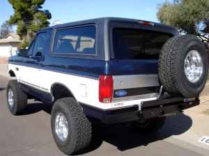 1993 Ford Bronco - Three-quarter rear view of a lifted dark-blue-over-silver full-size Ford Bronco with chrome modular wheels, oversized all-terrain tires, a…